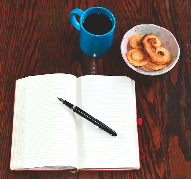 Notebook on a table with a black pen next to a cup of coffee and biscuits